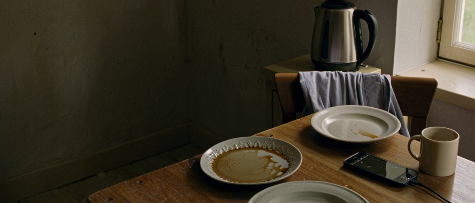a kitchen seen close with two plates left unwashed on the table, a phone charging beside a mug and a folded work blouse on the chair, the window and the kettle close behind