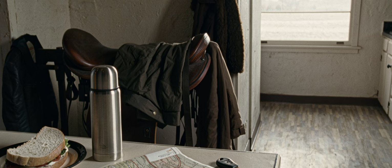 a ranch kitchen seen close with a truck key on the table beside a folded map with a long road traced in pen, a thermos and a plate with one sandwich, the saddle rack and the coats close behind