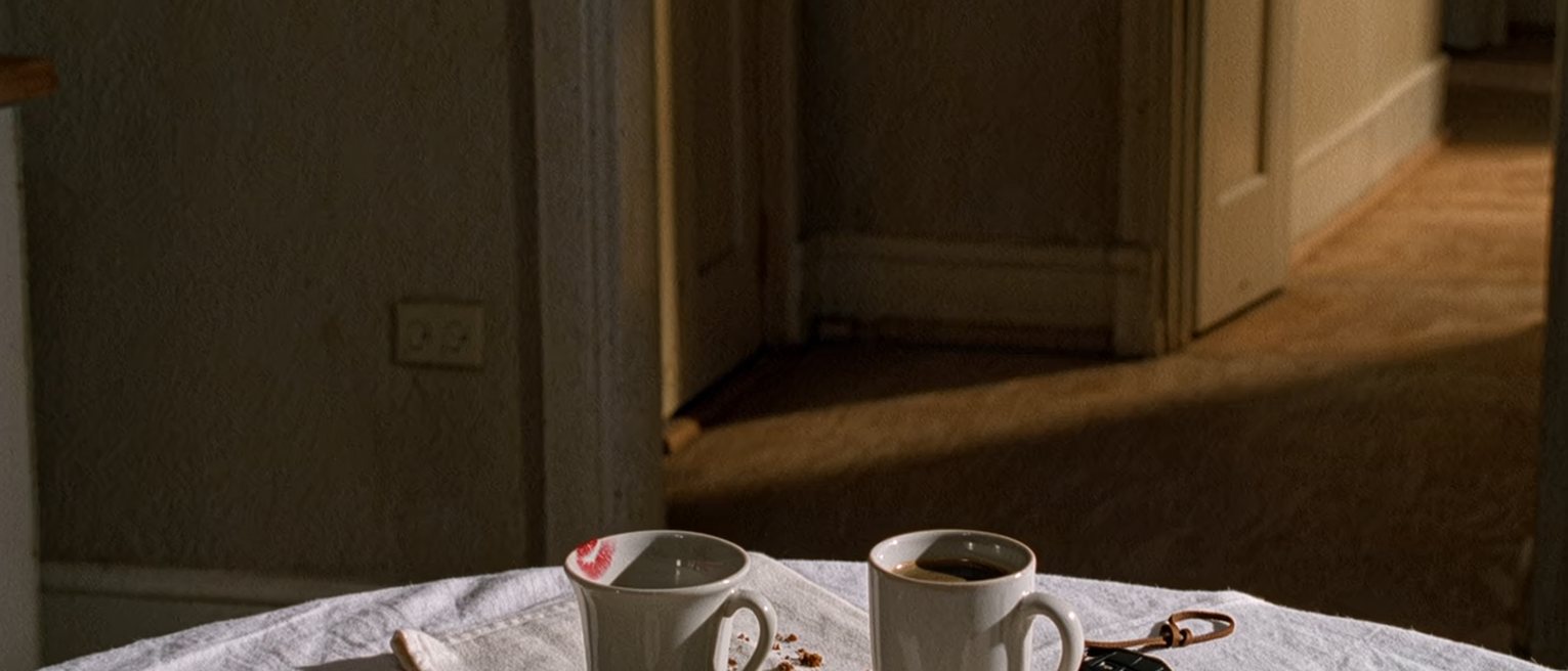 a kitchen table with two coffee cups, one with lipstick on the rim, and a set of car keys, crumbs still on the cloth