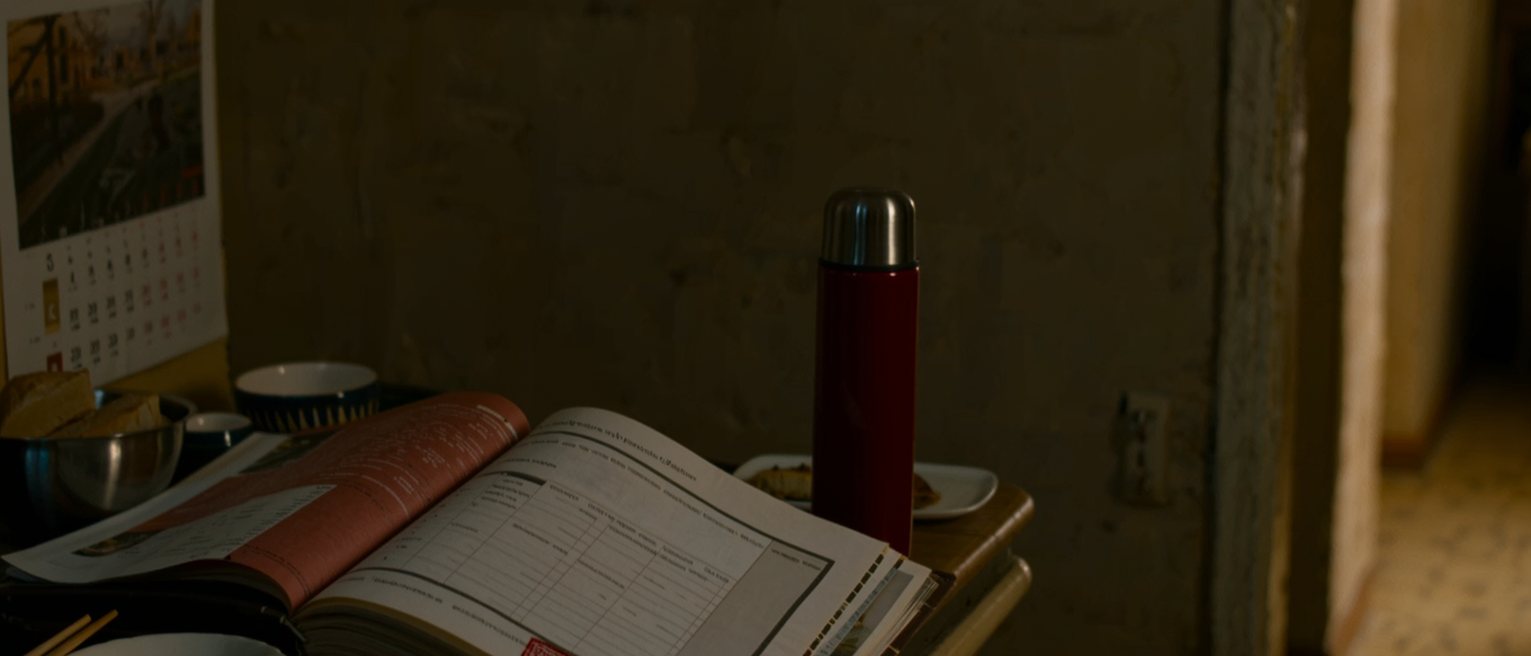 a kitchen table seen close with a family planning booklet open beside a printed permit stamped in red, a bowl of rice and a single pair of chopsticks, a red thermos and the wall calendar soft behind