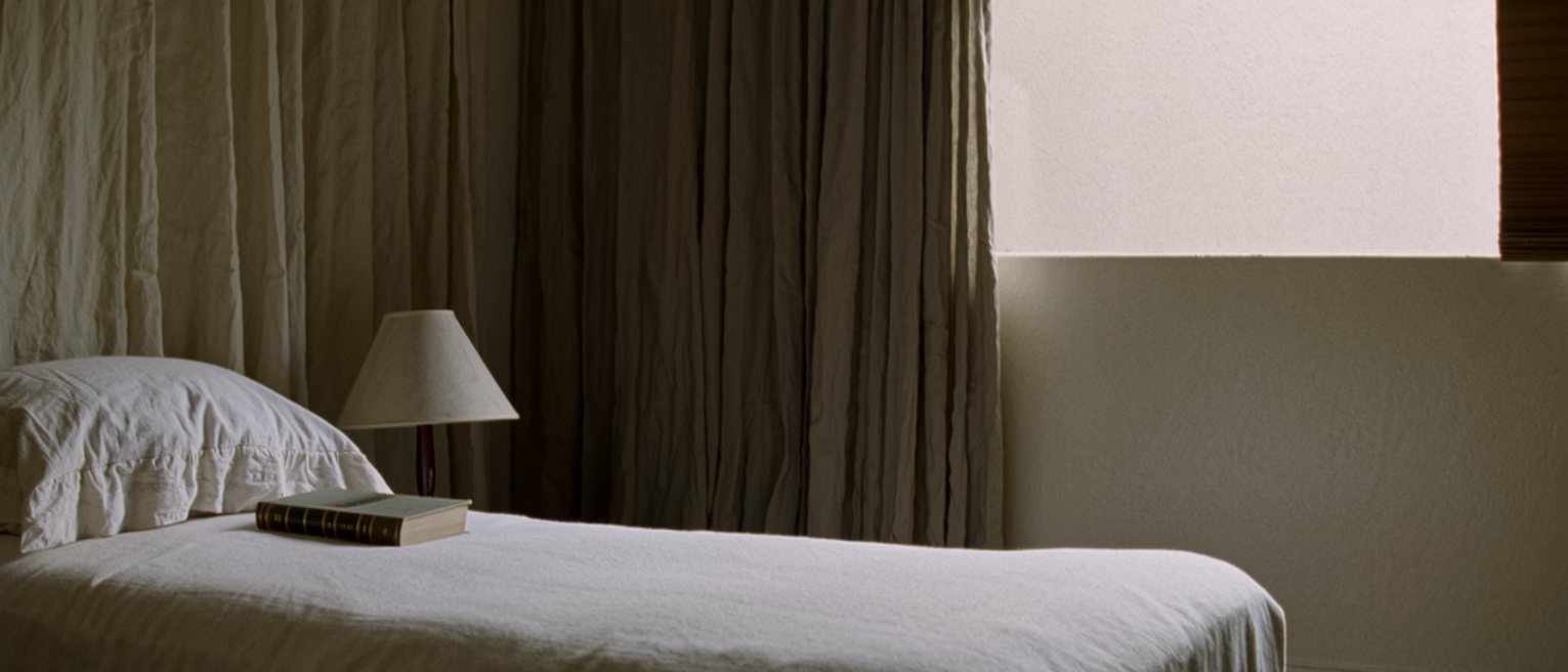 a bedroom seen close with the bed made and turned down on both sides, two books on the near pillow, a lamp with its shade tilted toward the bed and the curtains drawn behind