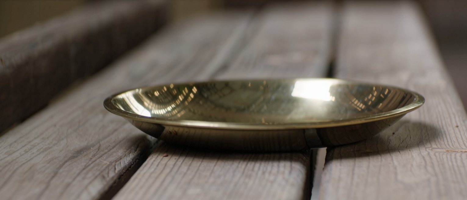a brass alms dish polished bright, lying open side up on a wooden bench