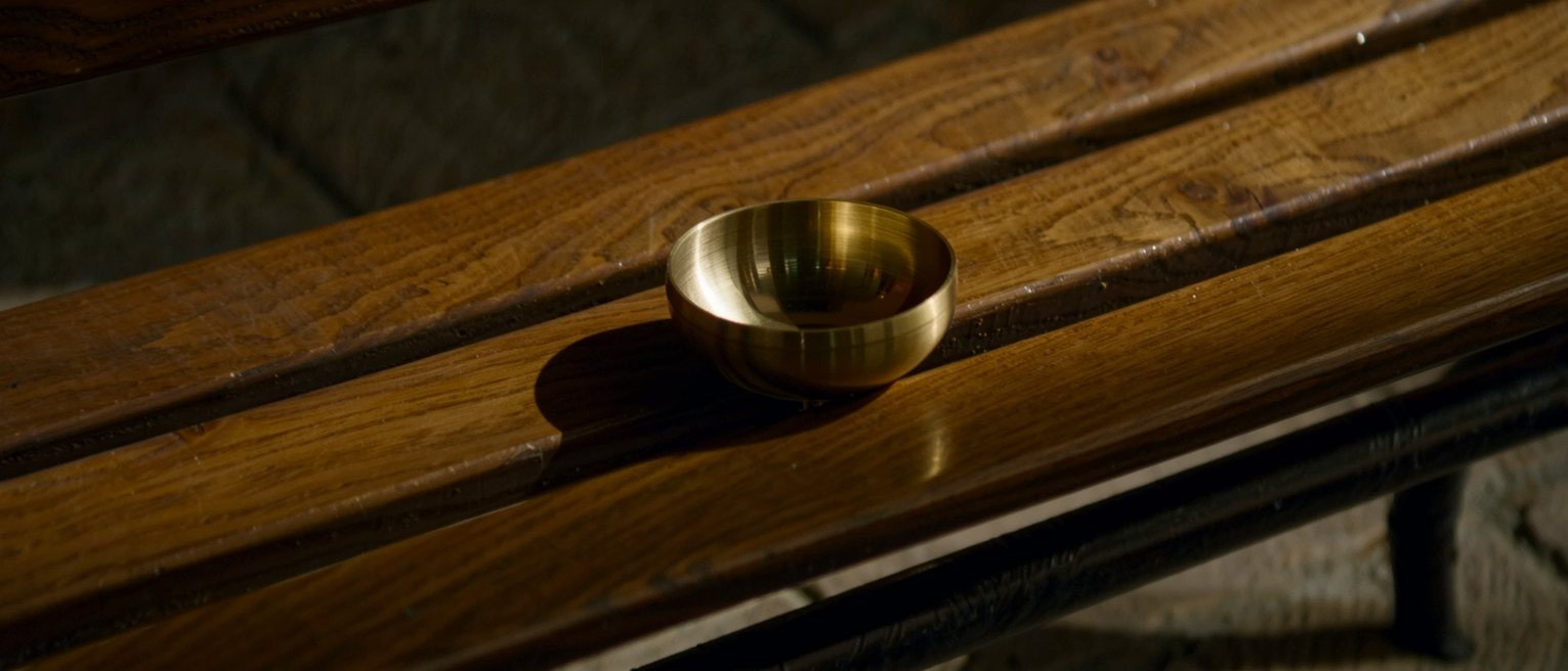 a brass alms dish polished bright, lying open side up on a wooden bench