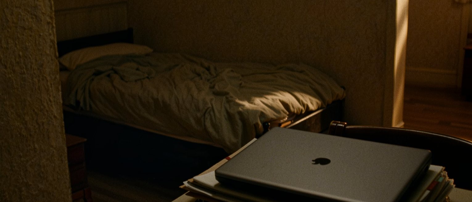a desk at home with a laptop closed on a neat stack of folders and a pen laid dead square to the edge, and beyond it a bed with the covers never turned down