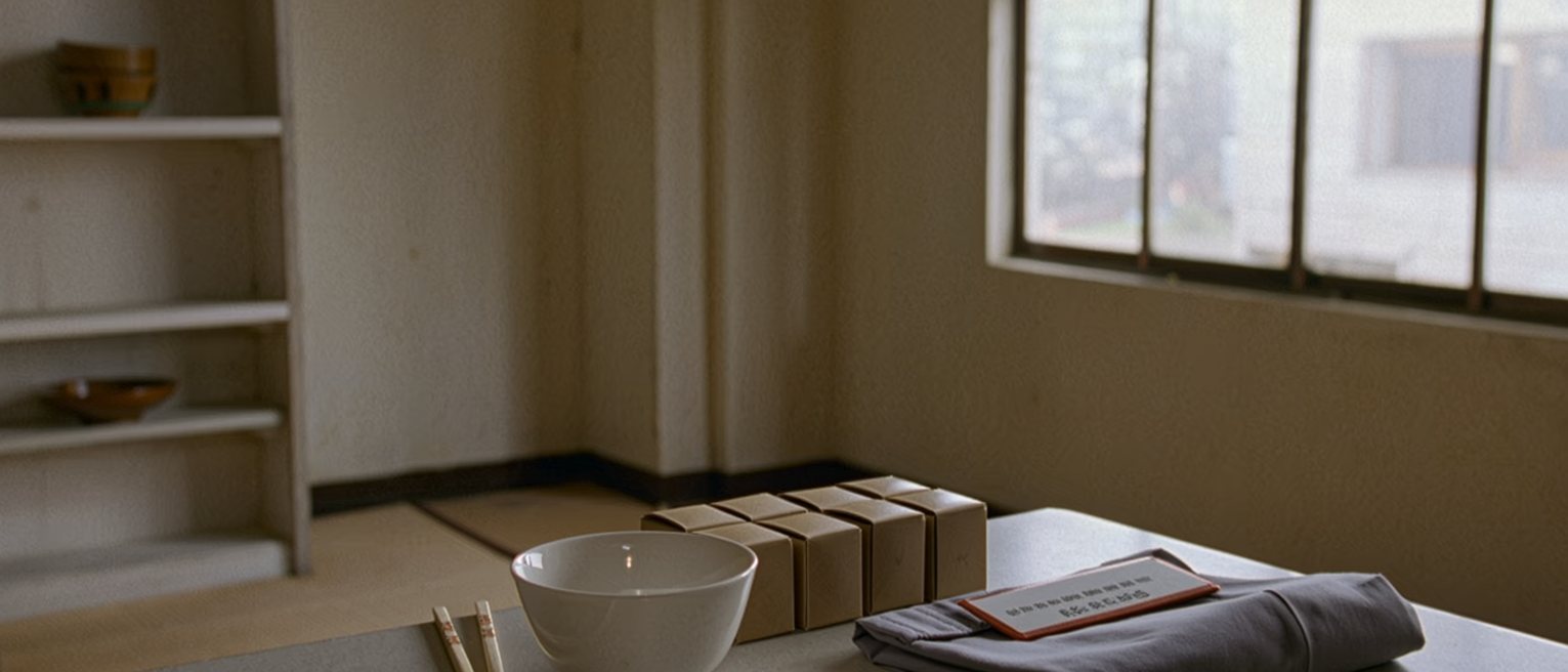 a small flat's kitchen seen close with a single bowl, chopsticks and a boxed meal laid out exactly square on the table, a name badge and a folded uniform shirt beside them, the shelves soft behind