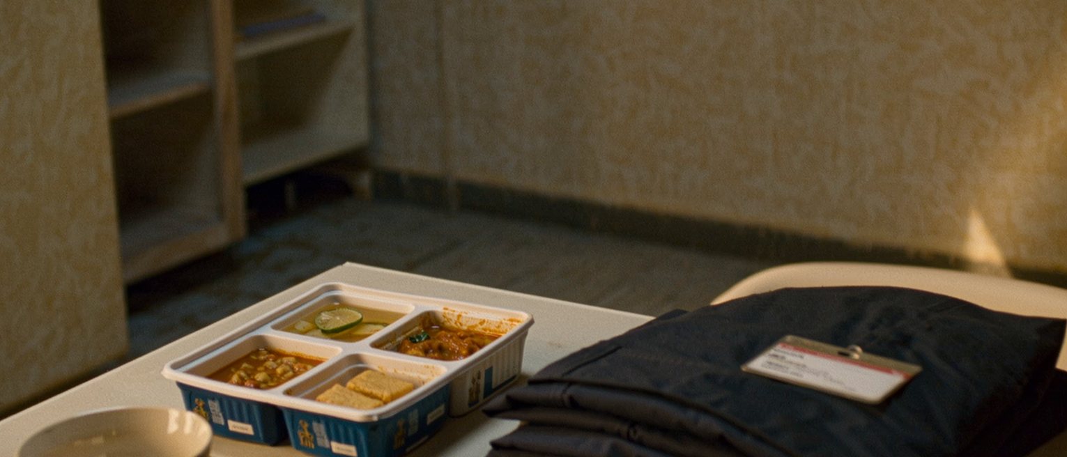 a small flat's kitchen seen close with a single bowl, chopsticks and a boxed meal laid out exactly square on the table, a name badge and a folded uniform shirt beside them, the shelves soft behind