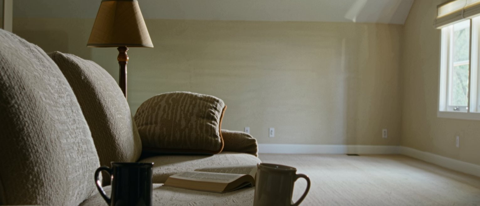 a sitting room seen close, two mugs on the low table at either end of the sofa, a book open face down between them, one cushion still dented, the lamp close behind