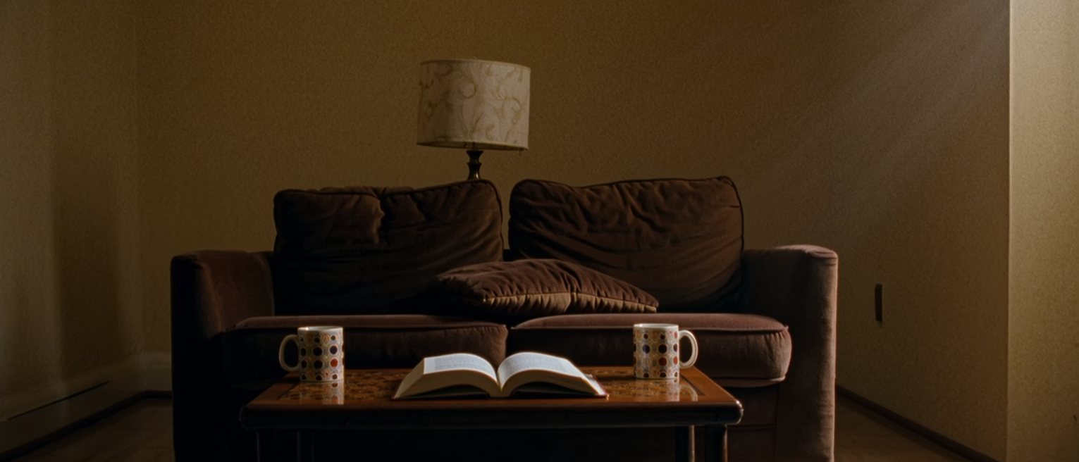 a sitting room seen close, two mugs on the low table at either end of the sofa, a book open face down between them, one cushion still dented, the lamp close behind