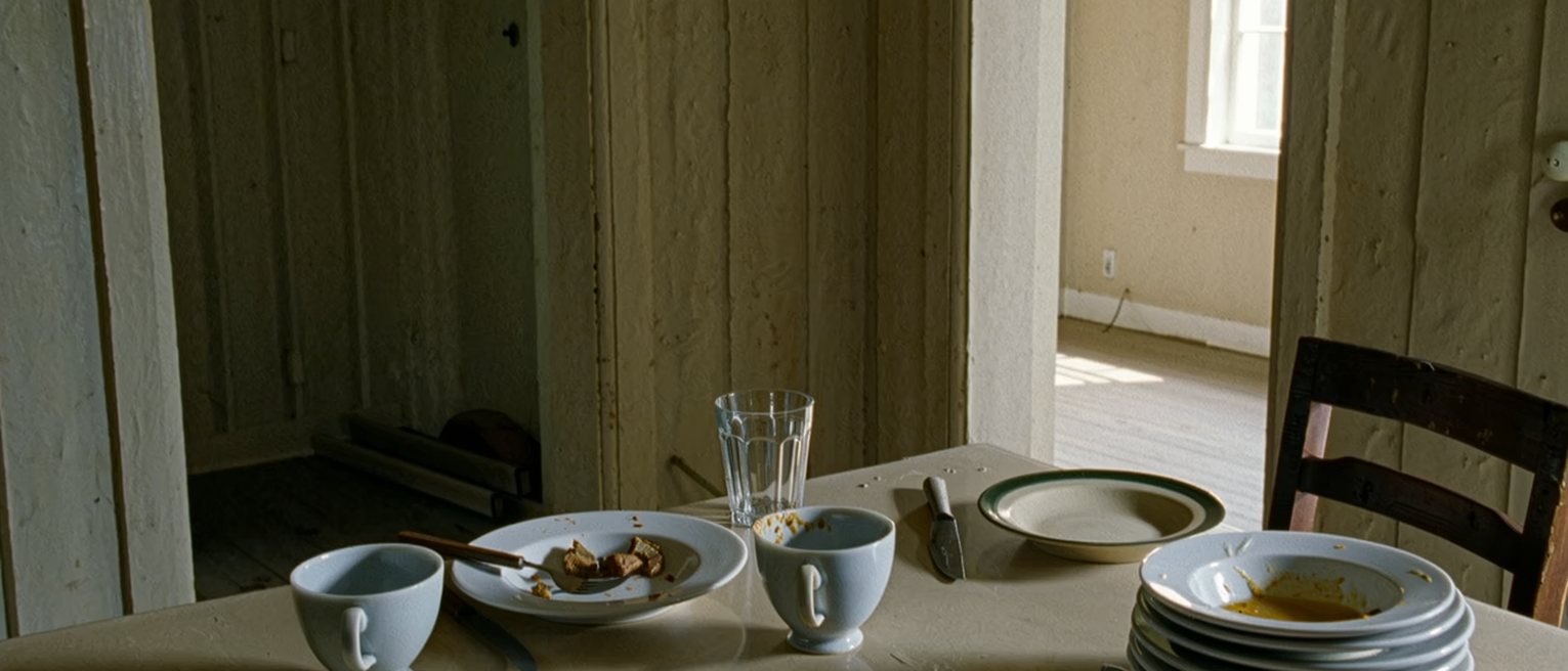 a kitchen with the breakfast things for four left unwashed for days, the table still laid for four, one chair pulled out