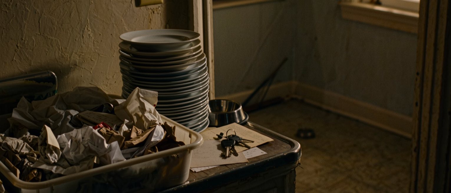 a kitchen seen close with the bin overflowing, a stack of unwashed plates in the sink, a dog bowl empty and a set of keys on the table beside an unopened letter, the window soft behind