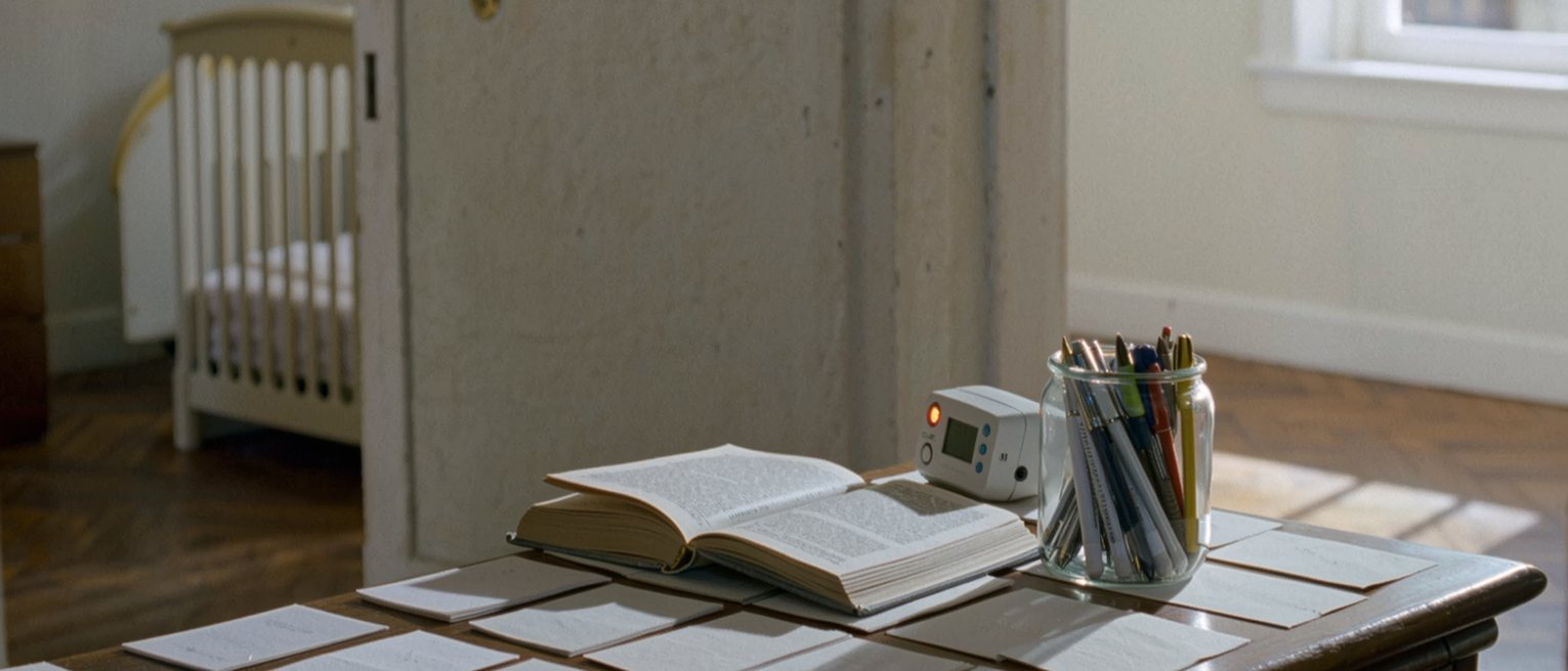 a desk seen close with index cards laid out in rows, a paperback of philosophy, a jar of pens and a baby monitor with its light on, the cot rail soft behind