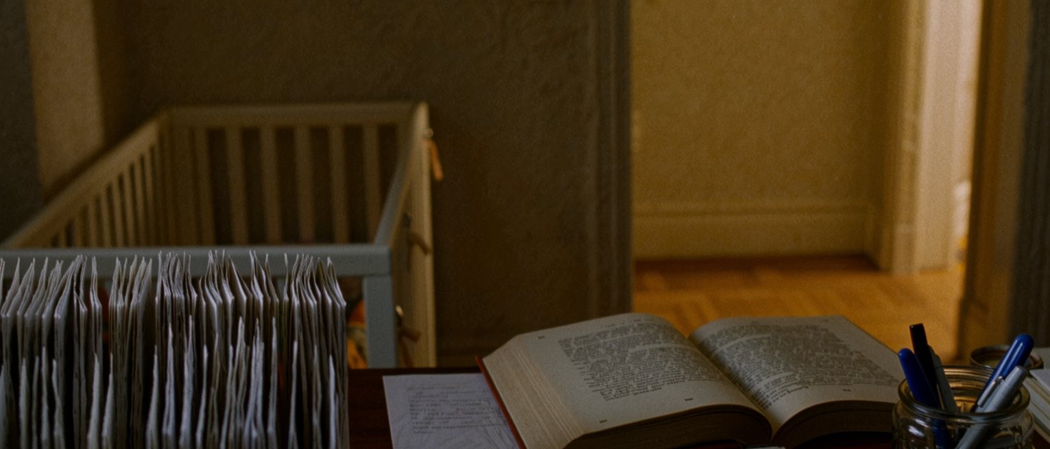 a desk seen close with index cards laid out in rows, a paperback of philosophy, a jar of pens and a baby monitor with its light on, the cot rail soft behind