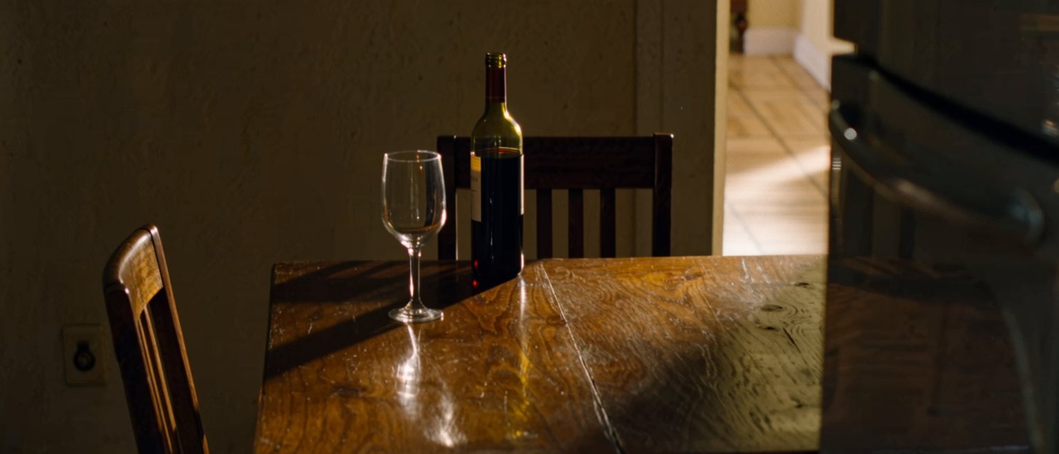 a kitchen table with a single cup, an open bottle of wine beside it and a second glass unpoured, the second chair pulled out and waiting