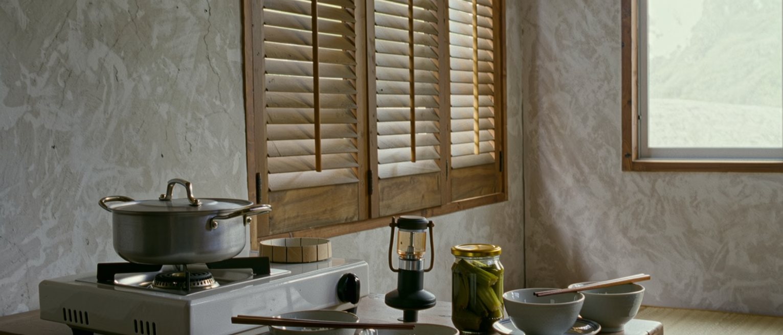 a mountain kitchen seen close with a pot of rice on the stove, three bowls and three pairs of chopsticks laid at one end of the table, a jar of pickles and a torch, the wooden shutters soft behind