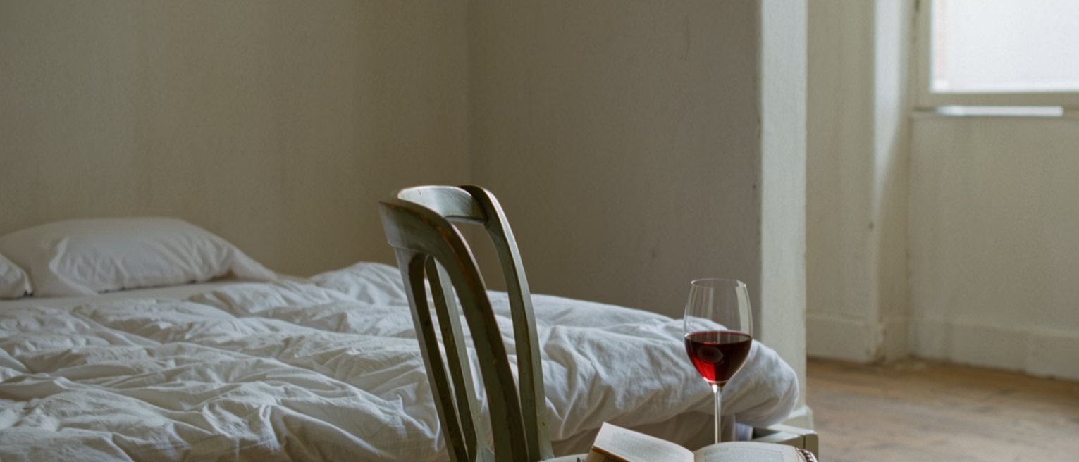 a bedroom with the covers still made and a single chair pulled to the bedside, a book left open on it and a glass of wine untouched