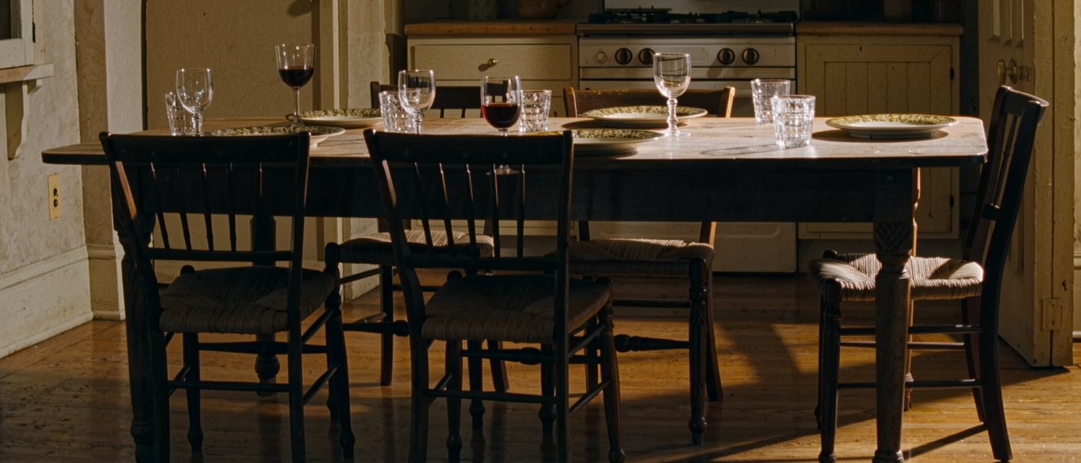 a kitchen table close in at the end of a long supper for six, plates and mismatched wine glasses still where they were set down, every chair still standing on the floor and turned out from the table at its own angle