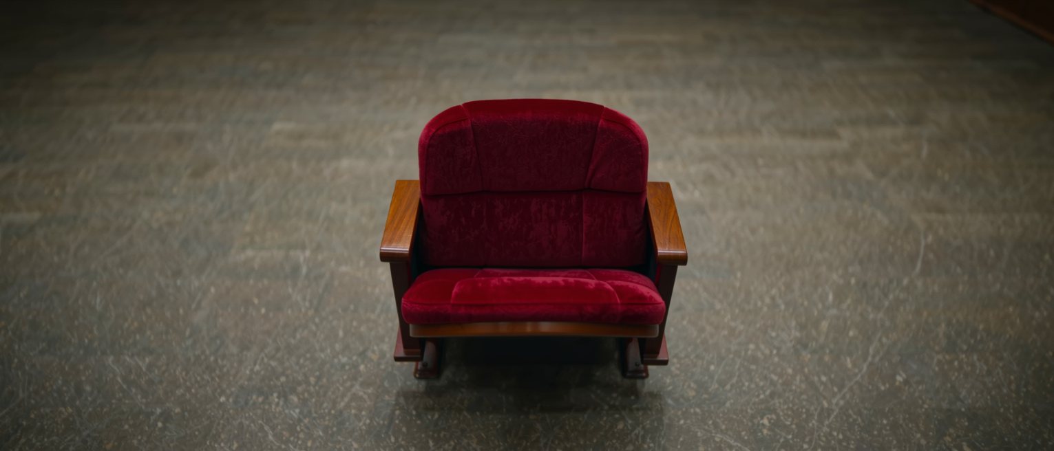 a single folded-up theatre seat of red plush with wooden armrests, standing alone at the end of a row of seats