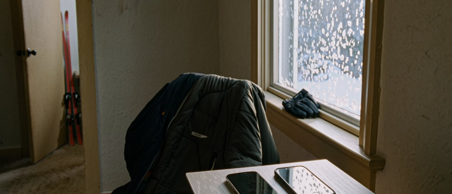 a bedroom seen close with two phones charging side by side on the bedside table, a ski jacket over the chair and a pair of gloves on the sill, snow on the glass behind