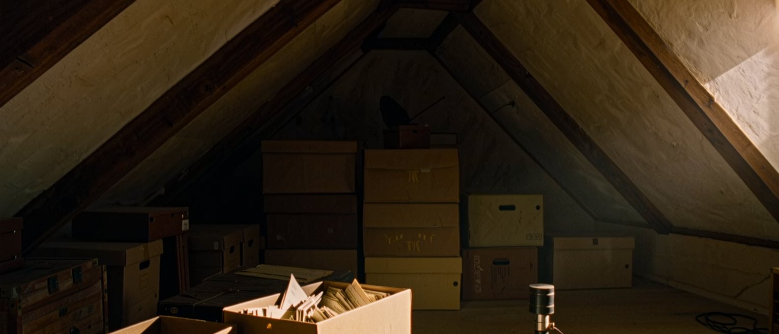 an attic seen close with a shoebox of old letters open on the floor, a slide viewer beside it and a torch, the rafters and a stack of boxes soft behind