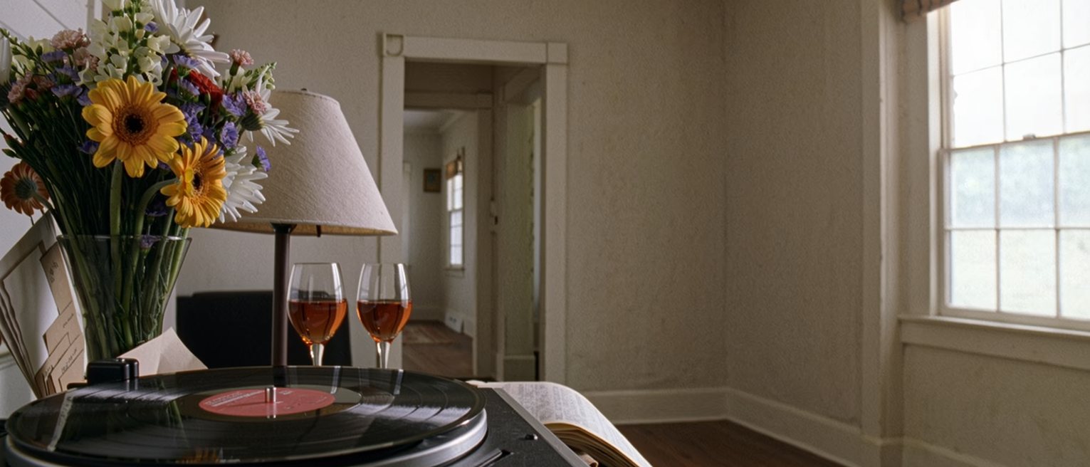 a sitting room seen close with a record on the turntable and a bouquet in its paper on the table, two glasses set out for a toast and a folded letter tucked under the vase, the lamp close behind