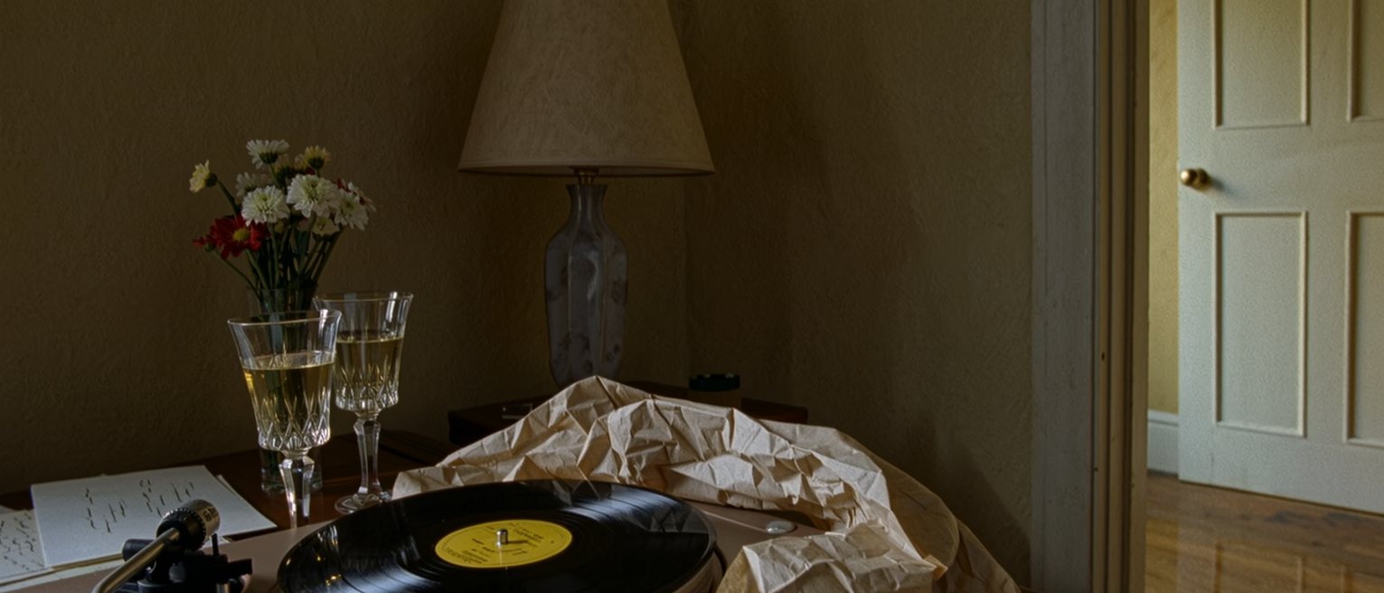 a sitting room seen close with a record on the turntable and a bouquet in its paper on the table, two glasses set out for a toast and a folded letter tucked under the vase, the lamp close behind