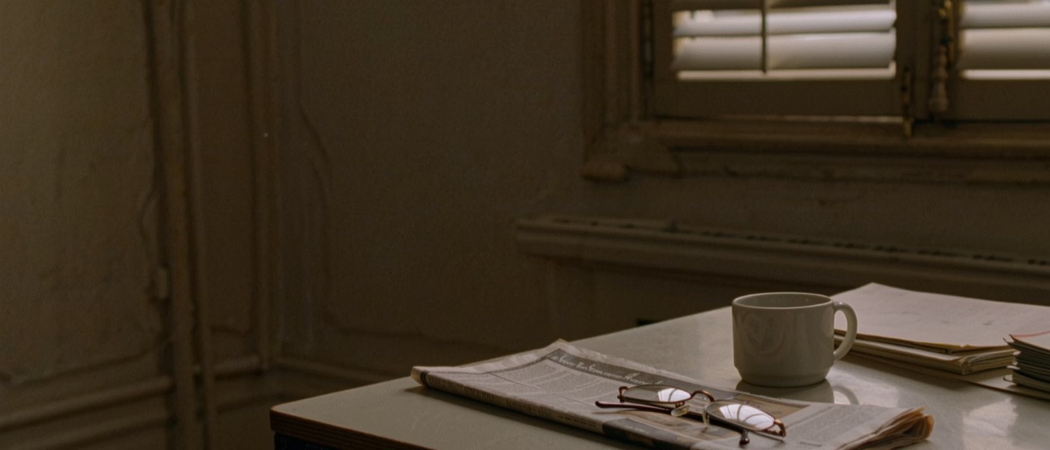 a Paris flat's desk seen close with a newspaper folded to a courtroom report, a pair of reading glasses on it, a coffee cup and a stack of paperbacks, the tall shuttered window soft behind