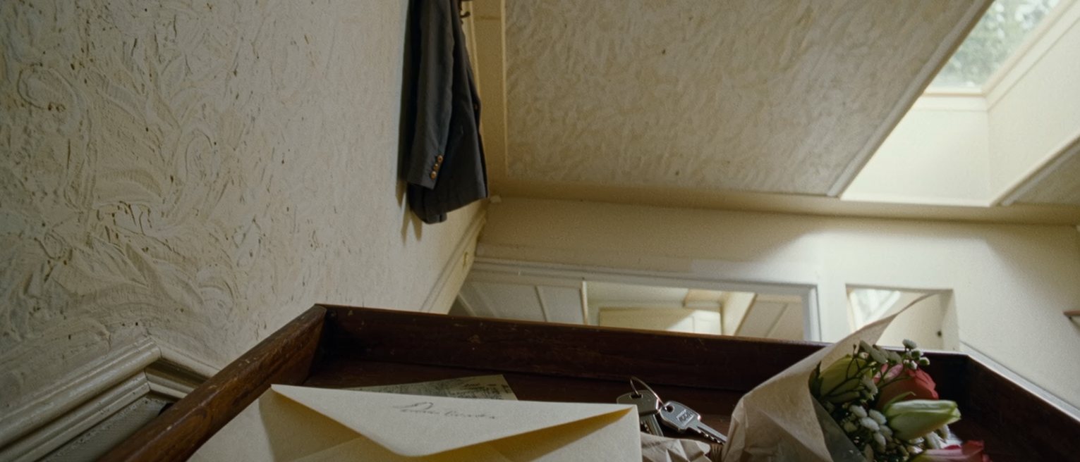 a hall console seen close with a letter in its opened envelope, a bunch of flowers still in their paper beside it, a set of keys, and a coat on the hook close behind