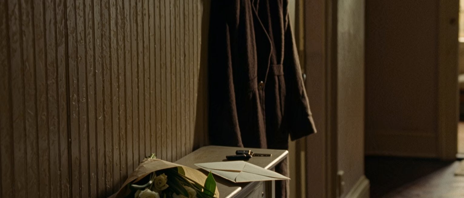 a hall console seen close with a letter in its opened envelope, a bunch of flowers still in their paper beside it, a set of keys, and a coat on the hook close behind