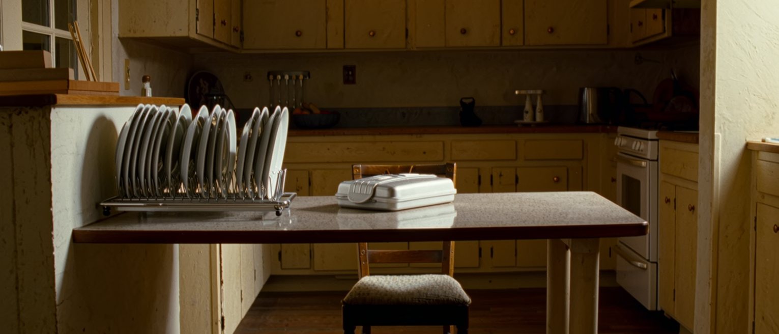 a kitchen after everything has been done: the counters wiped, the dishes racked, a lunchbox packed and closed, and one chair pulled out at the empty table