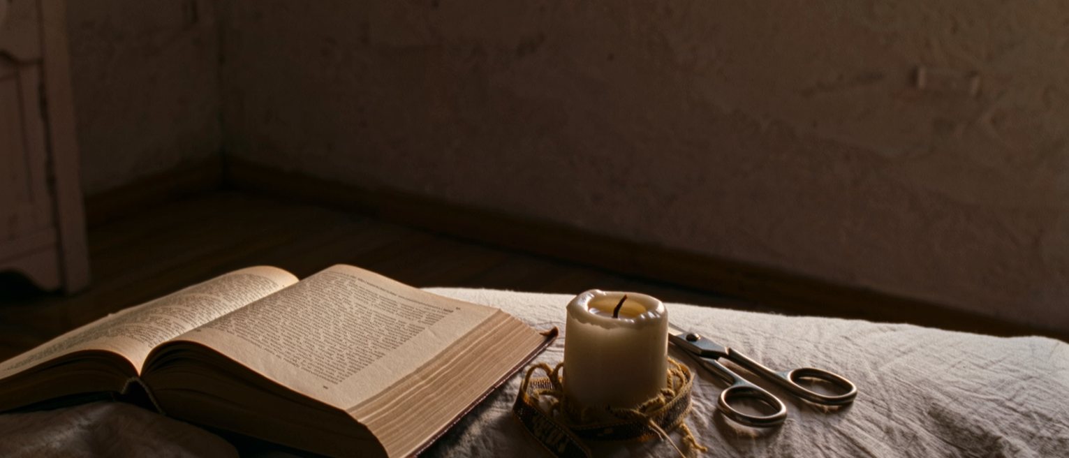 a bedroom with a book lying open and face down on the pillow, a candle burned down and a pair of scissors beside a cut ribbon