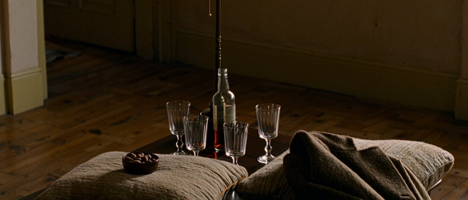 a low table seen close with four glasses and a bottle between four cushions, a bowl of nuts and a coat over one cushion, the lamp close behind