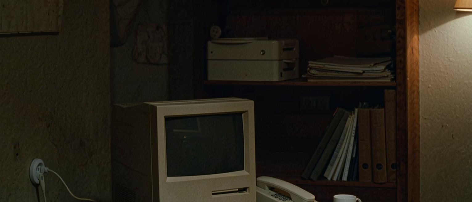a desk crowded close: a boxy beige computer with its monitor switched off, a keyboard, a phone cable trailing to the wall socket, a mug with a ring under it and a stack of papers beside the keyboard, the shelves behind soft