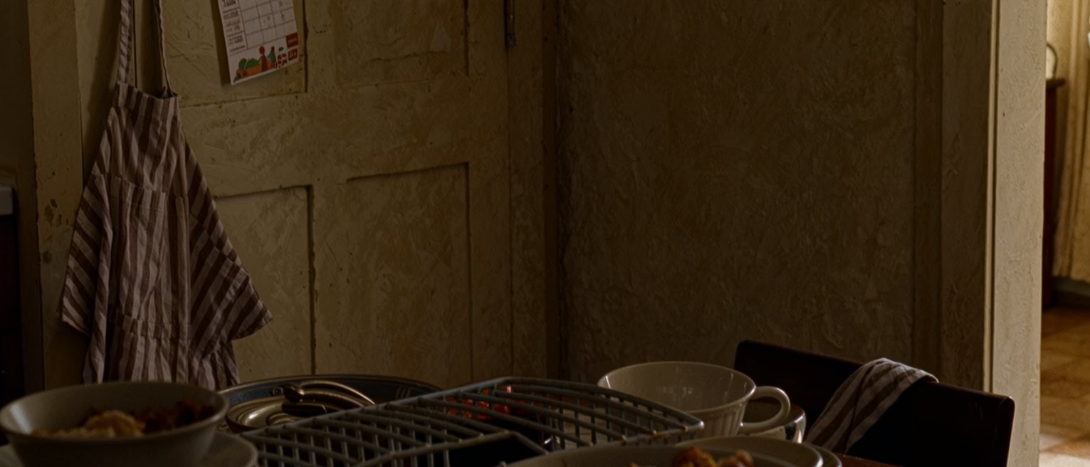 a kitchen counter seen close with the lunch things washed and racked, a child's timetable pinned to the fridge, an apron folded over the chair and a set of keys ready on the corner