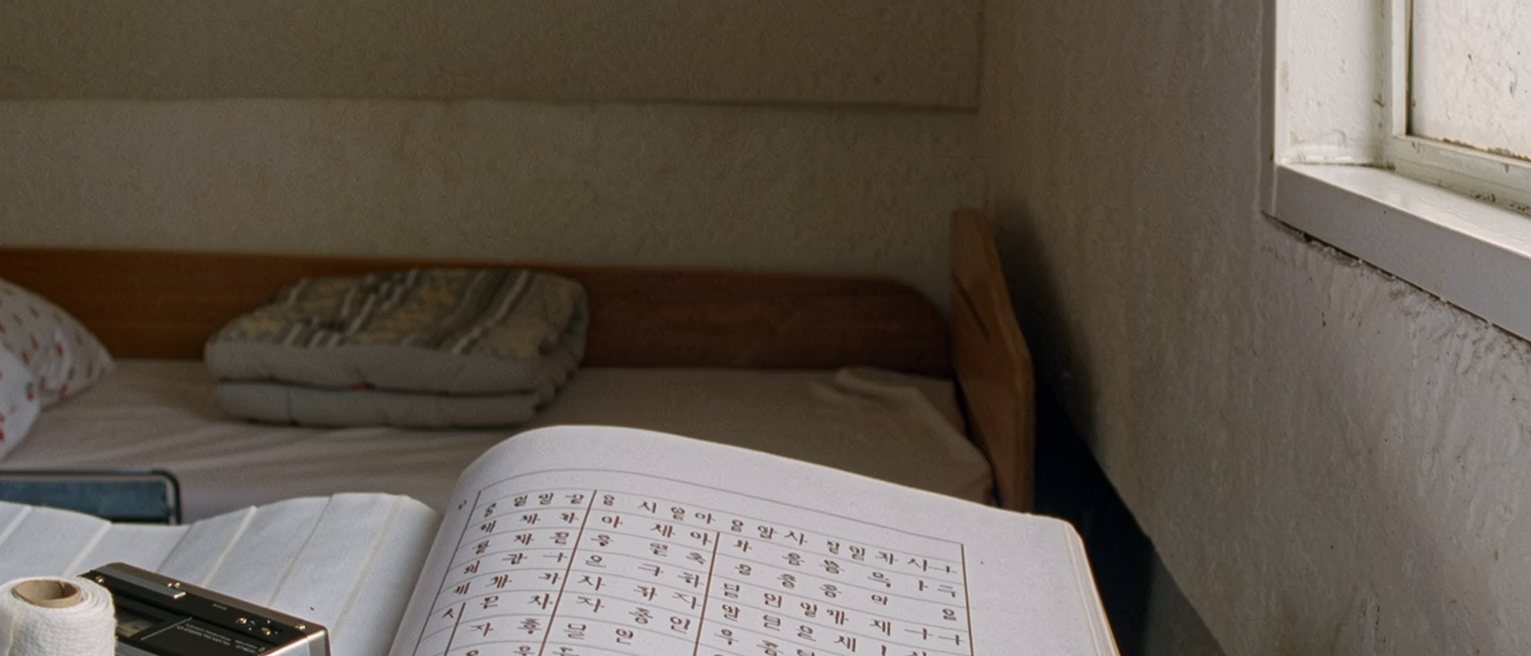 a small bedroom seen close with an exercise book open at a page of hangul practice, a cassette walkman, a roll of bandage and a rice cake wrapped in paper on the desk, the bunk bed soft behind