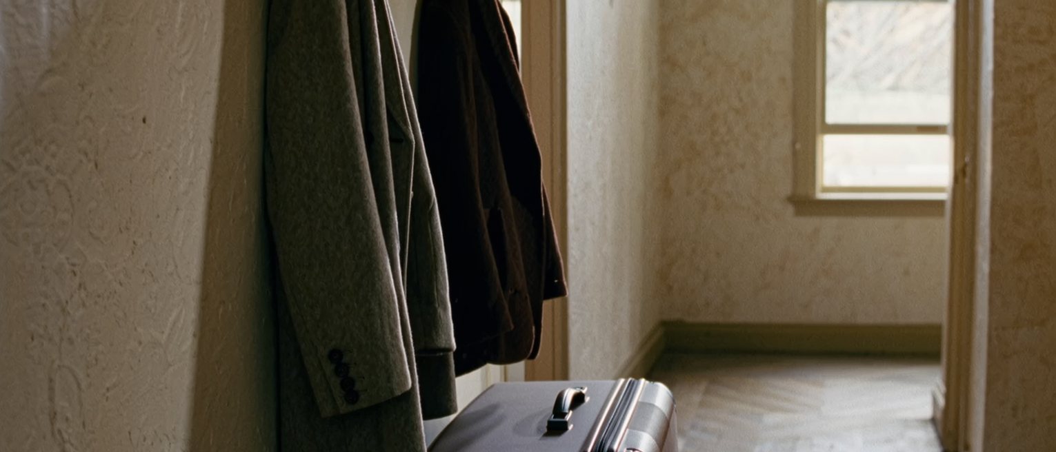 a hall with two coats on the hooks and, beneath them, one suitcase never unpacked from its last trip, a luggage label still tied to the handle