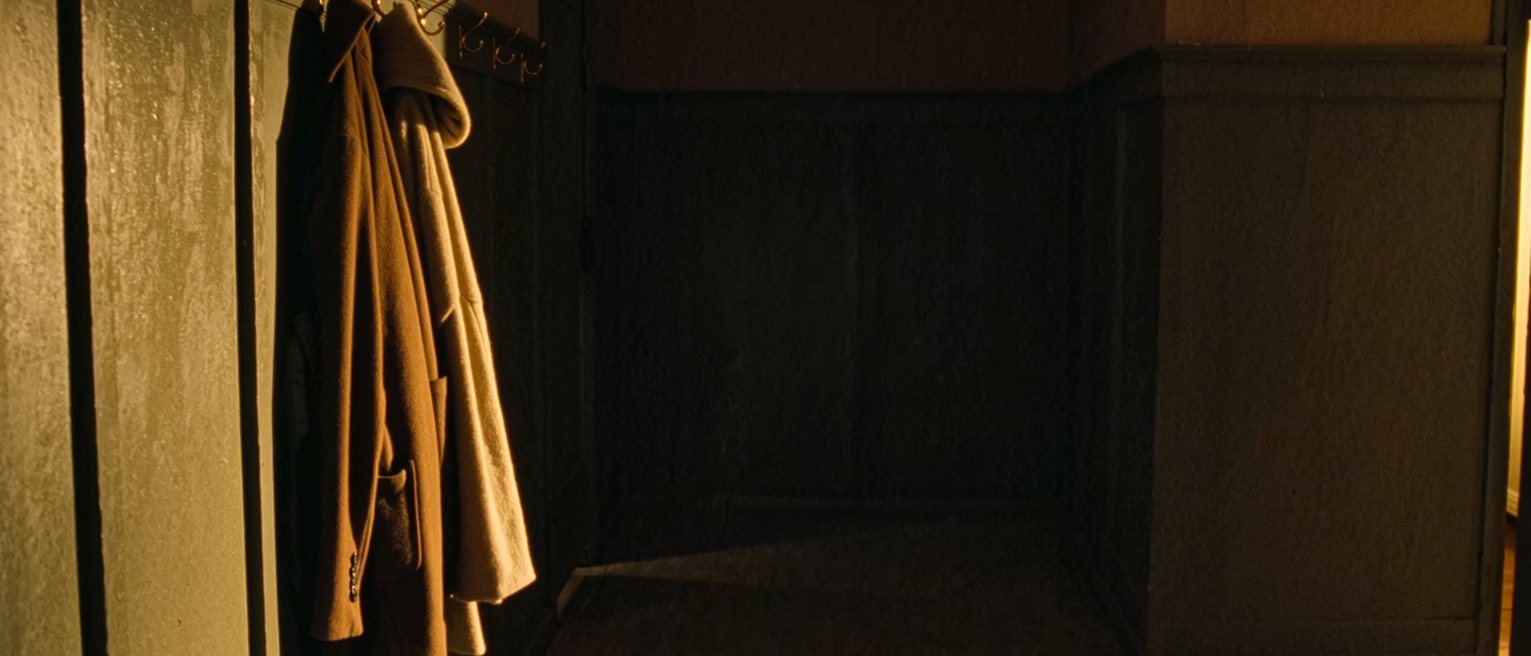 a hall with two coats on the hooks and, beneath them, one suitcase never unpacked from its last trip, a luggage label still tied to the handle