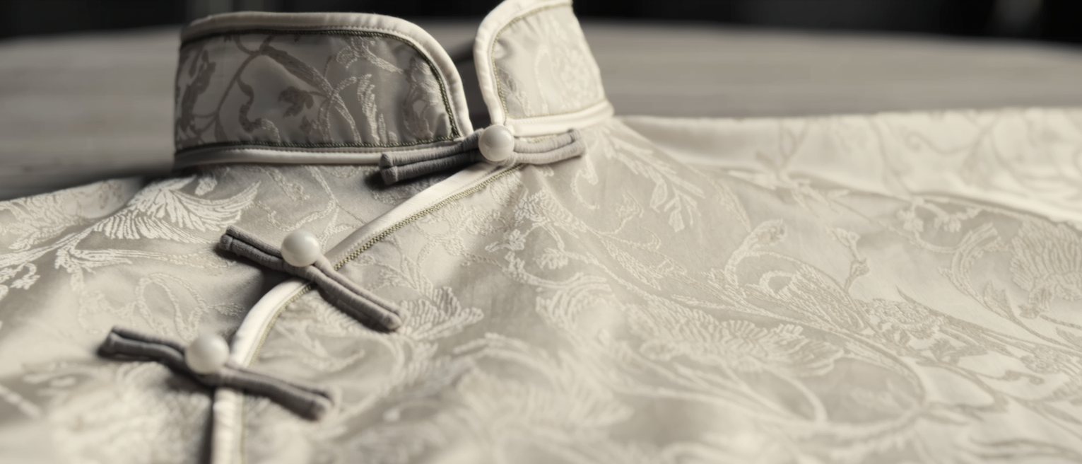 the high fastened collar of a patterned silk cheongsam lying empty and flat on a table, its cloth buttons closed one above another, no one wearing it