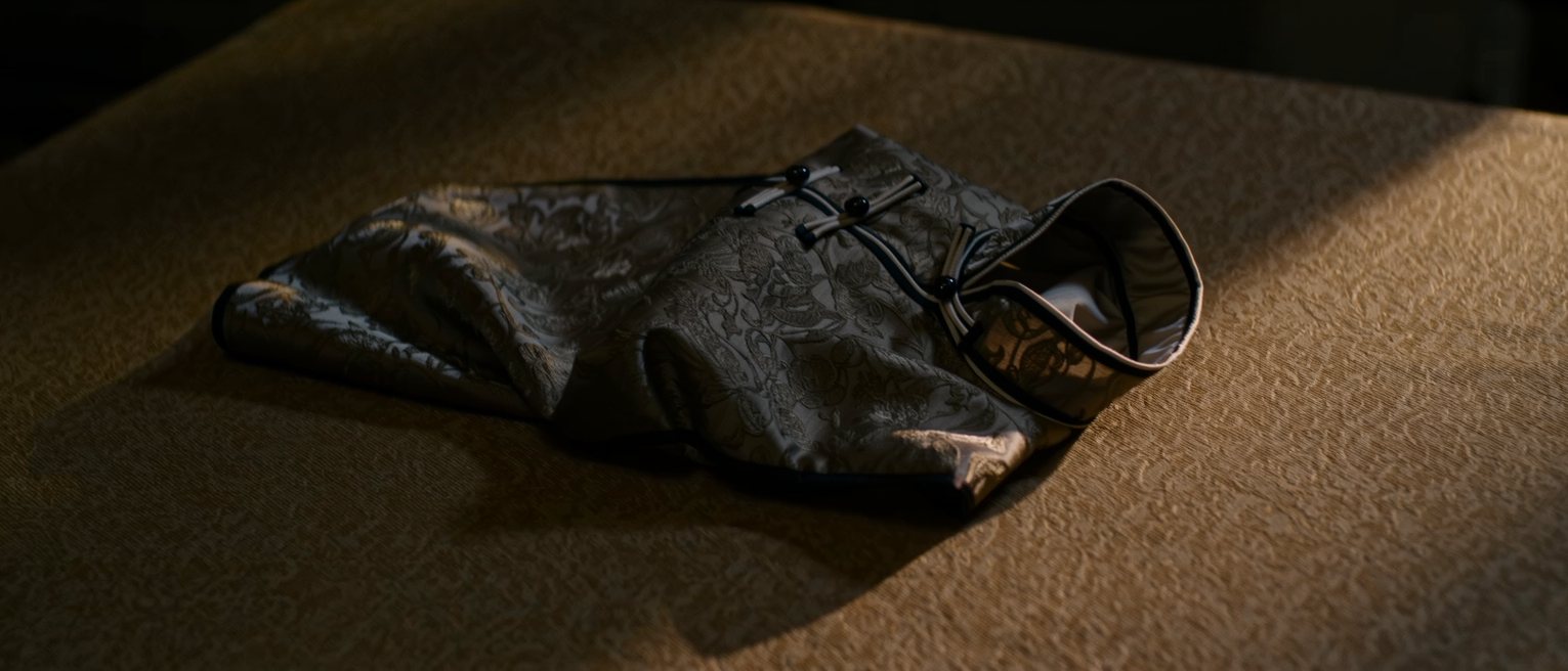 the high fastened collar of a patterned silk cheongsam lying empty and flat on a table, its cloth buttons closed one above another, no one wearing it