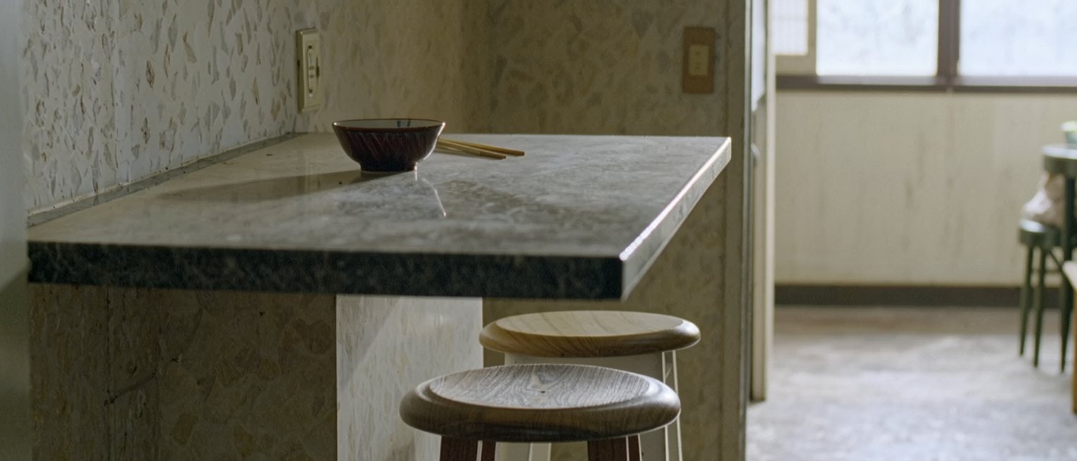 a narrow kitchen counter in a small flat with two stools, one pushed in and one left out, a cleared bowl and a pair of chopsticks standing at each