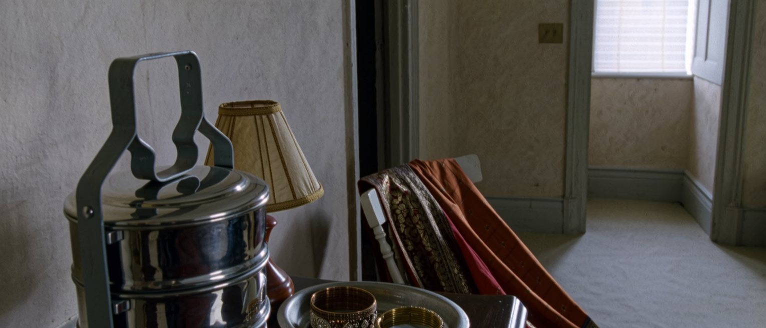 a bedroom seen close with a steel tiffin carrier on the dresser, a folded sari over the chair and two sets of bangles in a dish, the lamp close behind
