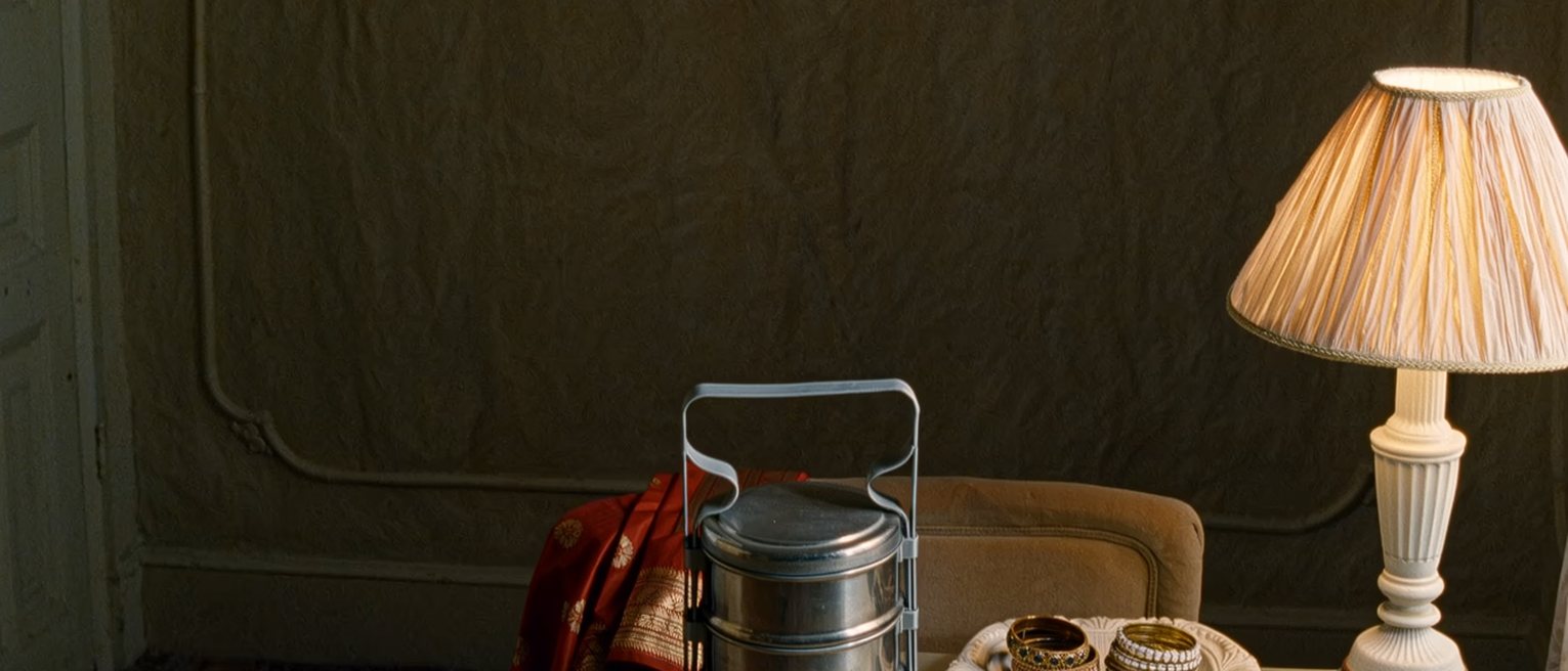 a bedroom seen close with a steel tiffin carrier on the dresser, a folded sari over the chair and two sets of bangles in a dish, the lamp close behind