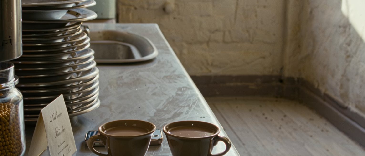 a kitchen seen close with two cups of chai, a wedding card propped against the jar of dal and a phone face down, the steel plates racked behind