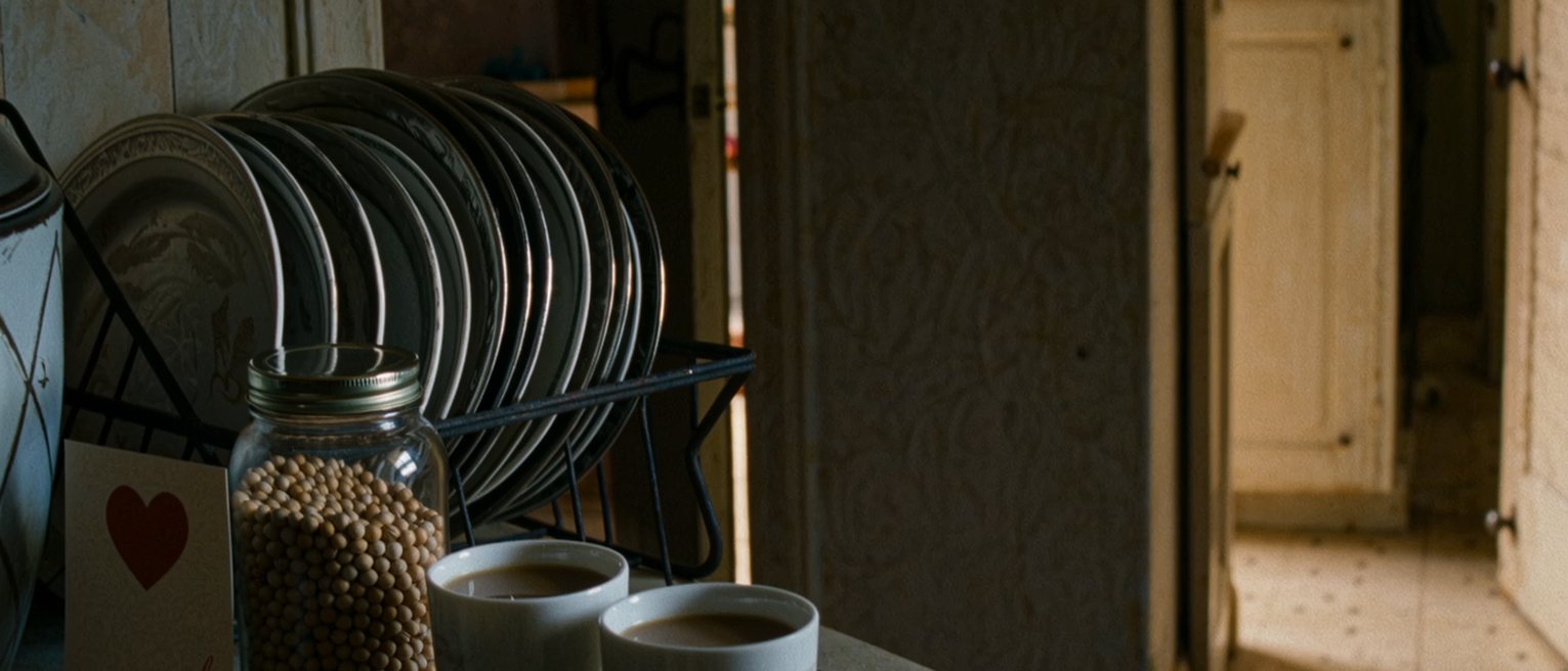 a kitchen seen close with two cups of chai, a wedding card propped against the jar of dal and a phone face down, the steel plates racked behind