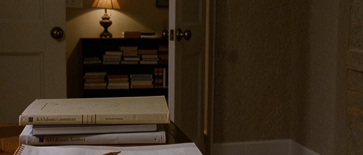 a desk seen close with two dictionaries closed and stacked, a blank notepad in front of them with a pencil laid across it, a lamp and a shelf of dictionaries soft behind