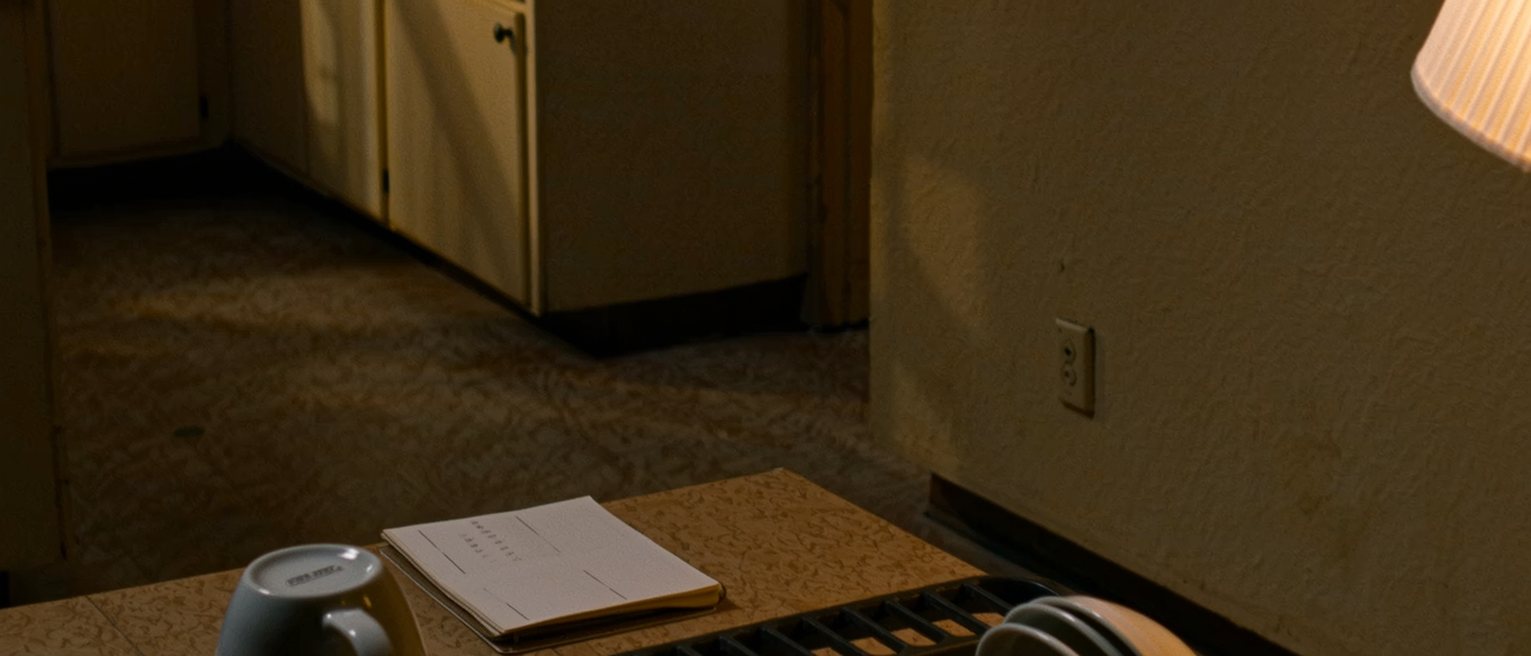 a kitchen table seen close with a notepad on it and nothing written, a single cup and a single plate washed and upturned on the rack close behind, the kettle and the window beyond