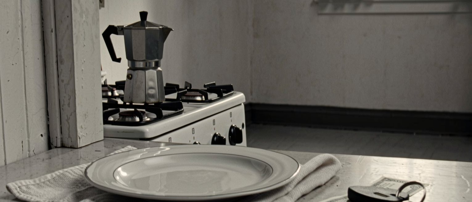 a kitchen table seen close with a plate kept warm under a cloth, a set of house keys and a pay slip folded beside it, the moka pot on the stove behind