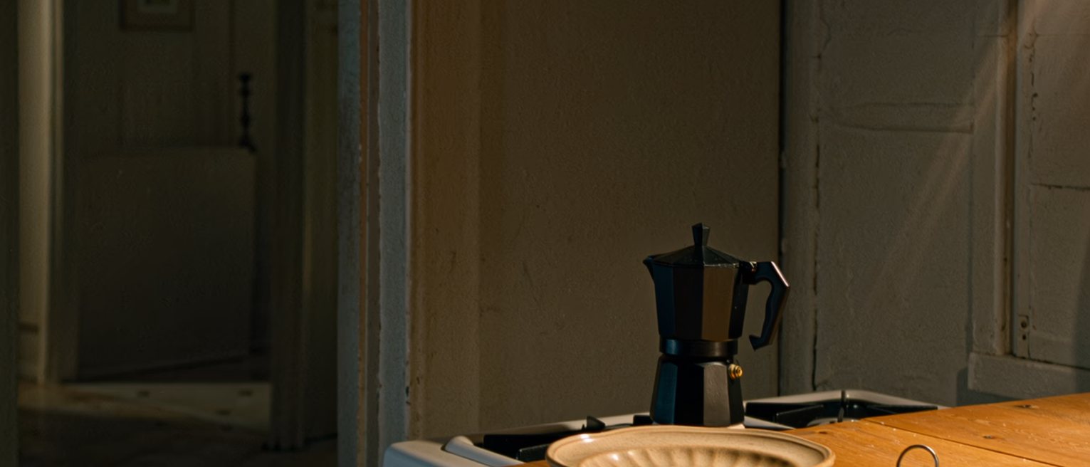 a kitchen table seen close with a plate kept warm under a cloth, a set of house keys and a pay slip folded beside it, the moka pot on the stove behind