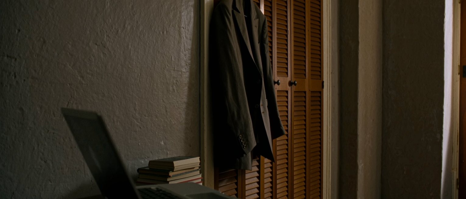 a bedroom seen close in a room kept as it was, a laptop on the desk beside a stack of textbooks, a suit on the wardrobe door and the shutters soft behind