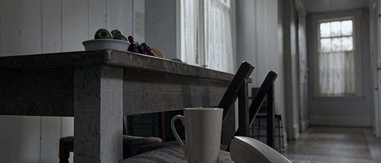 a kitchen table seen close with a cup gone cold beside a small flat handset lying face down, a chair pulled out at an angle, a bowl of fruit at the far end, the door soft beyond