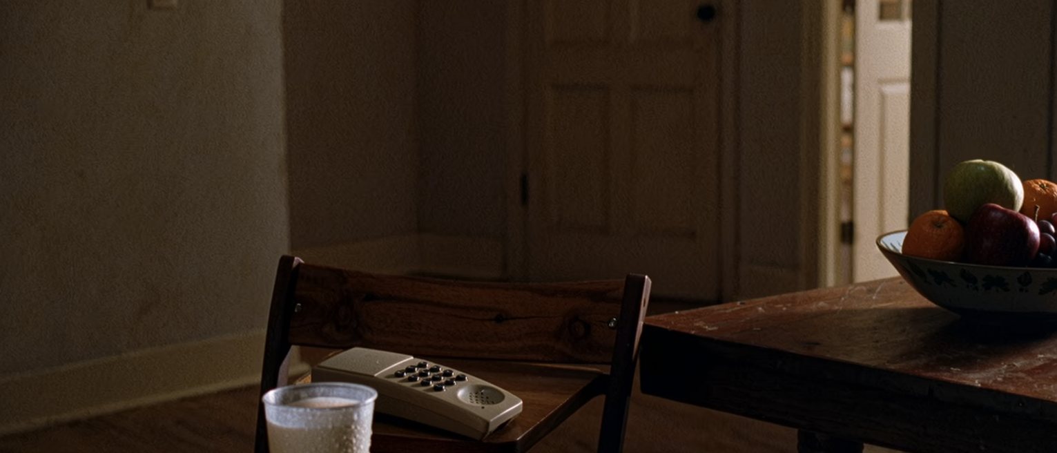 a kitchen table seen close with a cup gone cold beside a small flat handset lying face down, a chair pulled out at an angle, a bowl of fruit at the far end, the door soft beyond